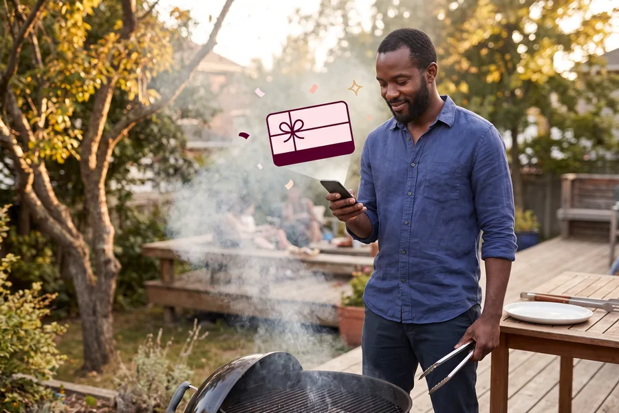 Man at a backyard grill with tongs down, reading a gift link on his phone