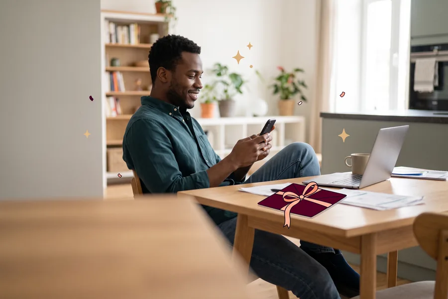 Man leaning back at his kitchen table, smiling at his phone beside a laptop