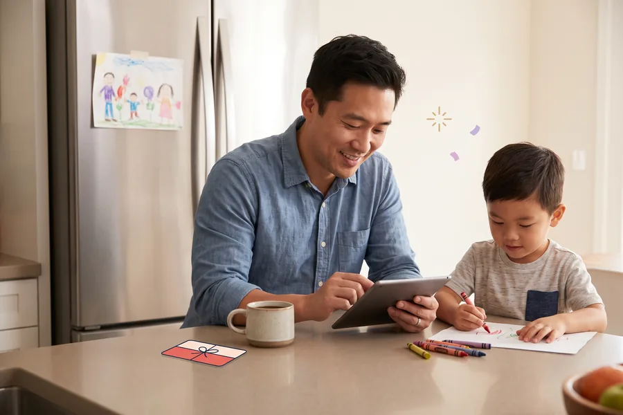 Dad at the kitchen counter scrolling a tablet while his young son draws with crayons