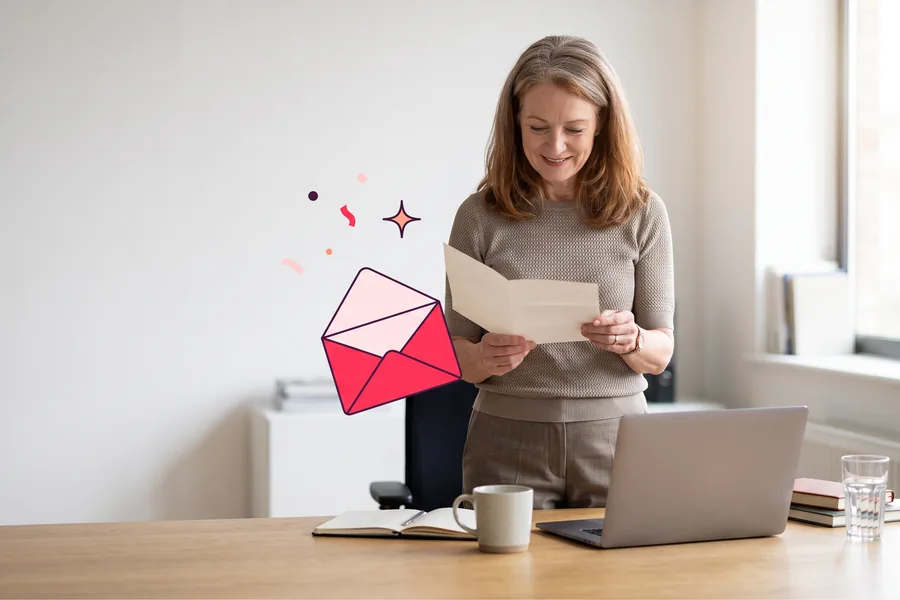 An Irish woman in her late fifties stands at a desk in a bright office, reading a folded paper note with a small smile in soft afternoon light