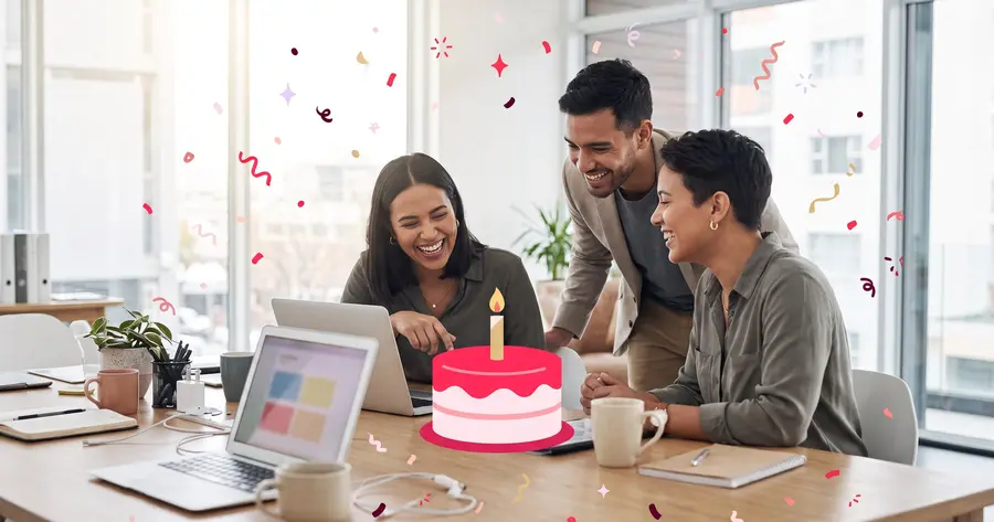 Coworkers laughing together at a desk as a teammate reads a birthday message on her laptop