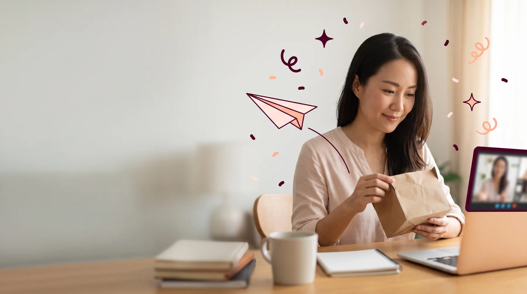 Remote employee at a home desk holding a just-opened parcel, a video call on her laptop