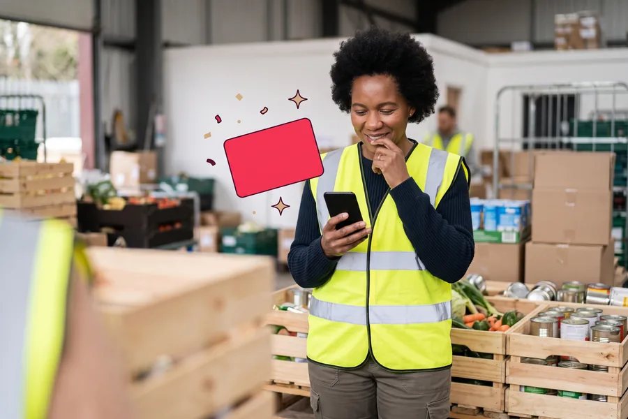 Volunteer in a hi-vis vest among crates at a food drive, smiling at her phone