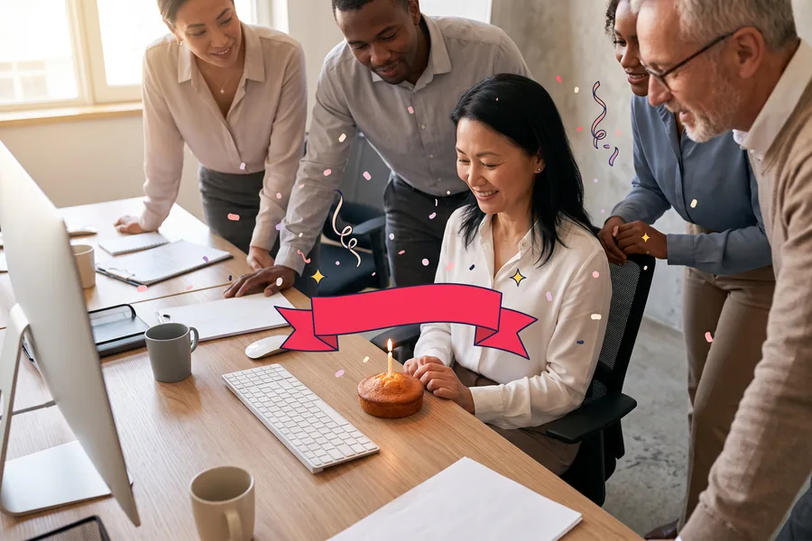 Colleagues leaning in around a coworker at her desk, a small cake with one candle by her keyboard