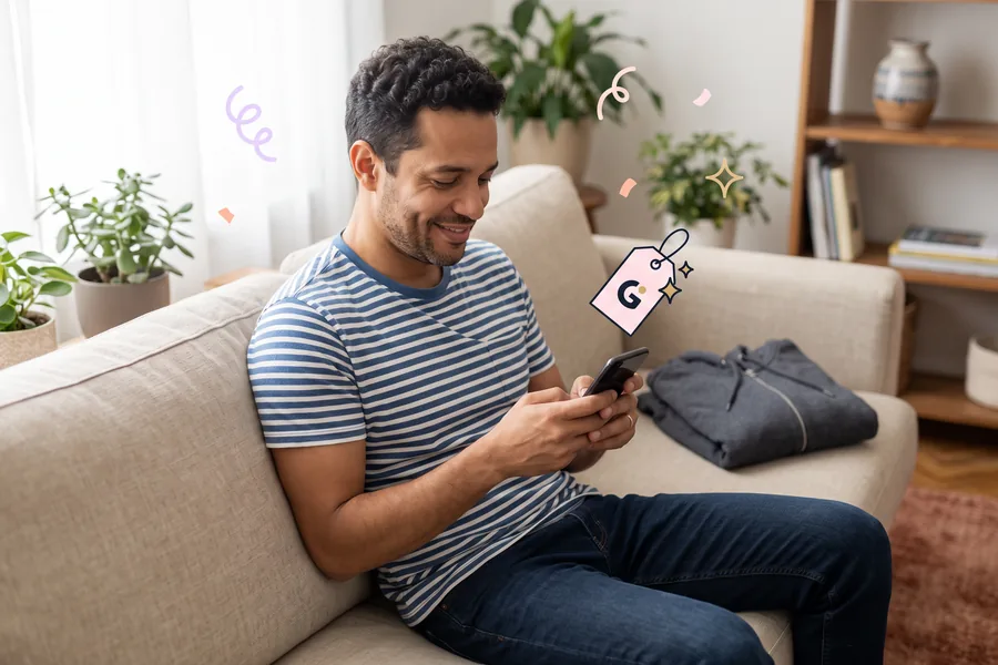 Man on a couch with a folded hoodie beside him, smiling at his phone